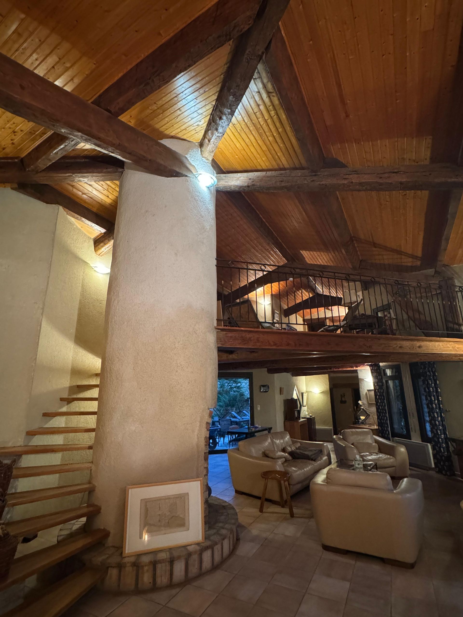 Vue intérieure : Plafond à poutres en bois, escalier en colimaçon, colonne en pierre, coin salon avec canapés.