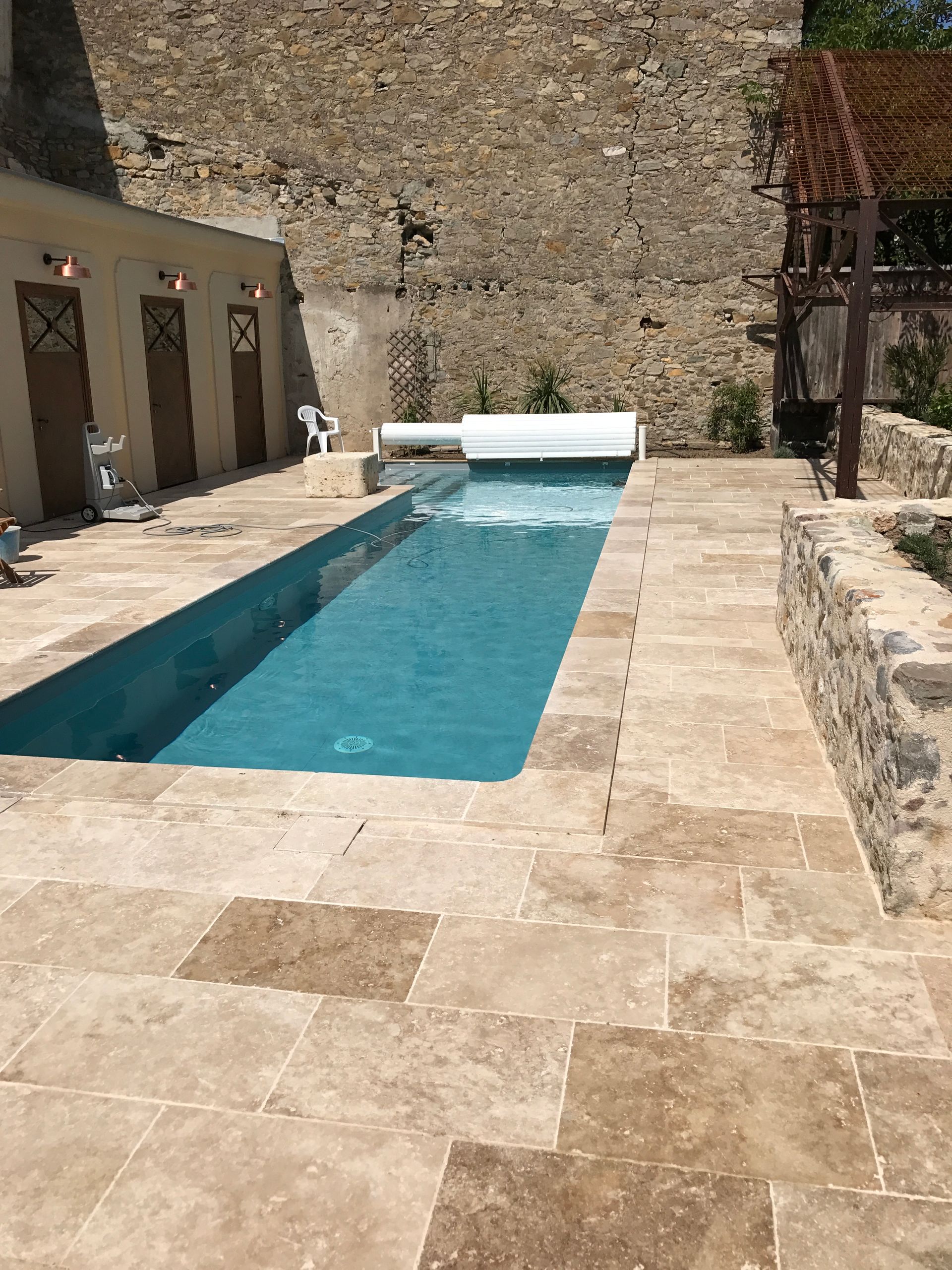 Piscine rectangulaire, entourée de dalles en pierre, à côté d'un mur en pierre et de petits bâtiments.