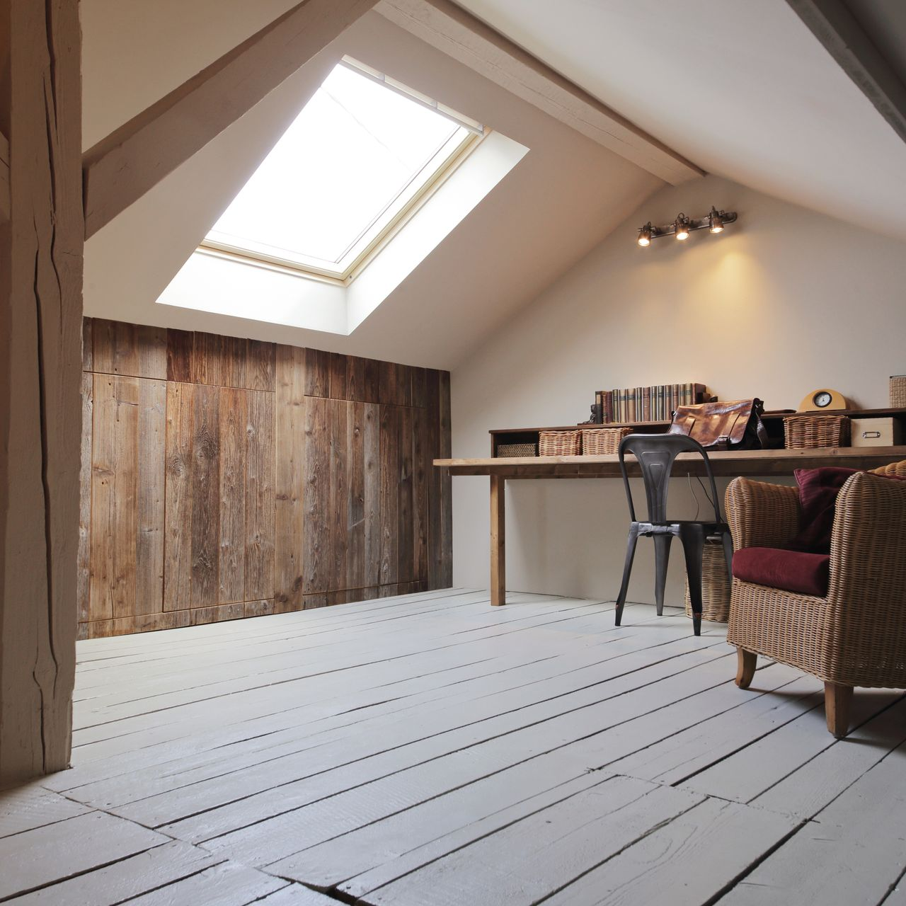 Chambre mansardée avec mur en panneaux de bois, bureau, puits de lumière, chaise en osier et poutres en bois naturel.