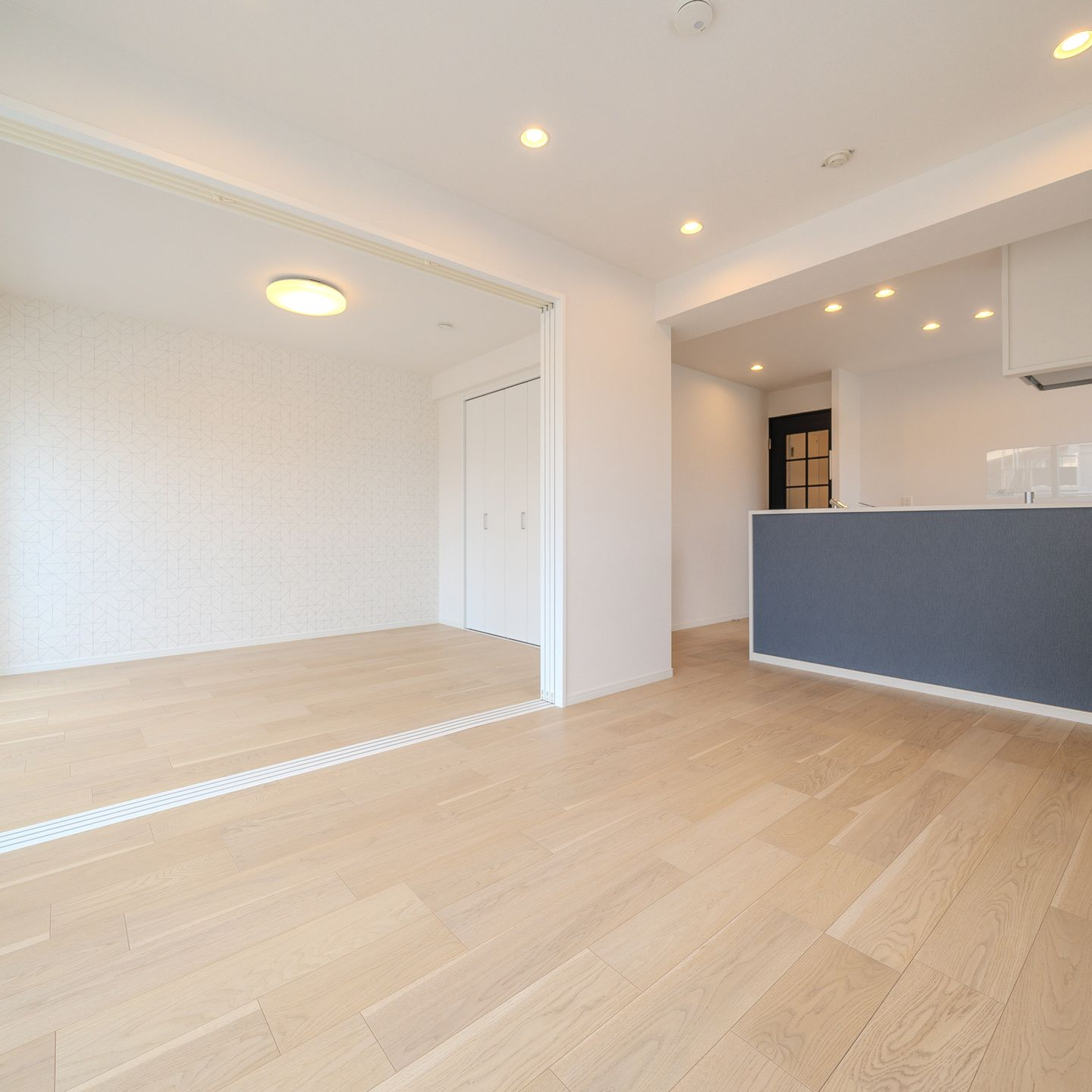 Pièce vide et lumineuse avec parquet en bois clair, une cuisine en arrière-plan et une porte coulissante.