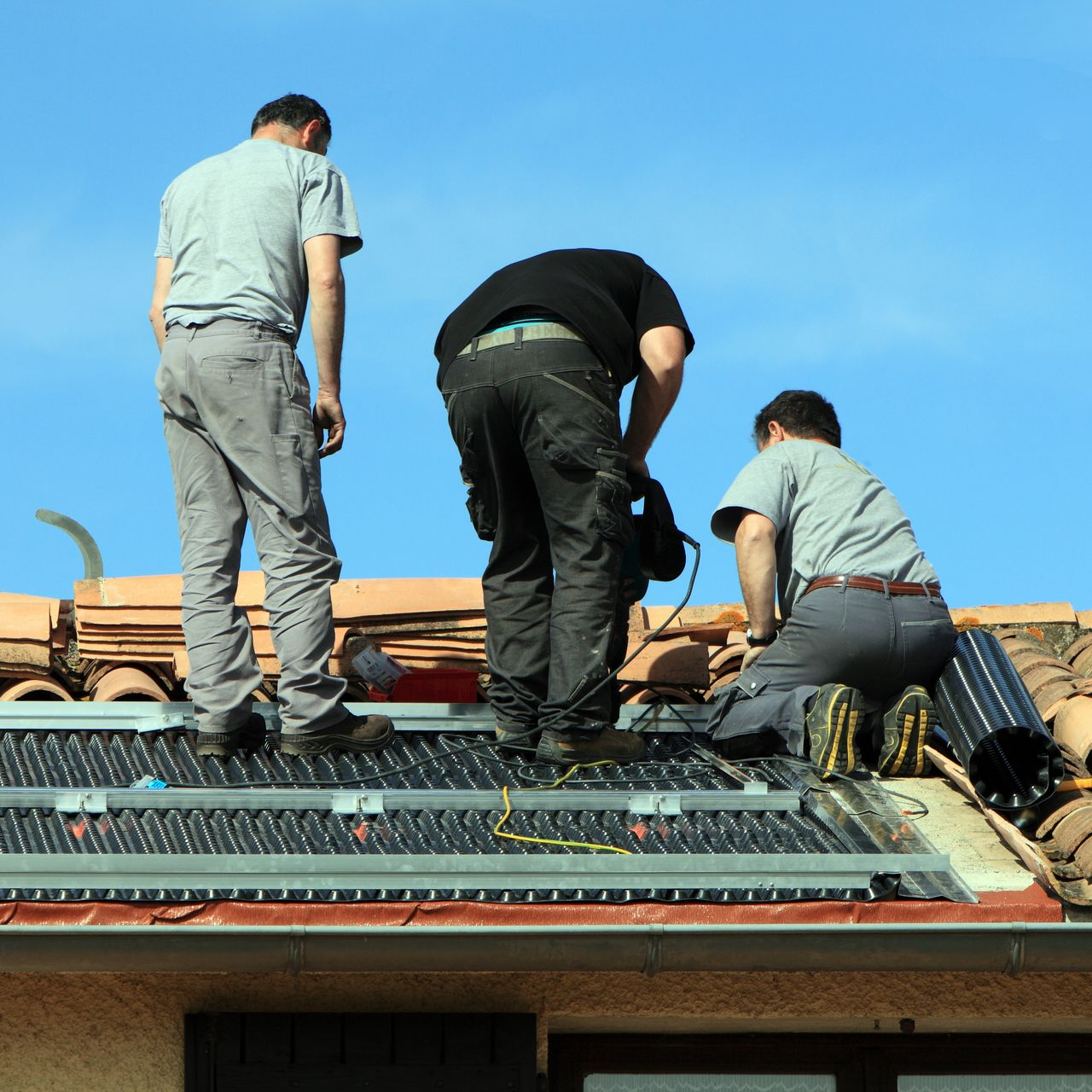Trois ouvriers installent des panneaux solaires sur un toit sous un ciel bleu.