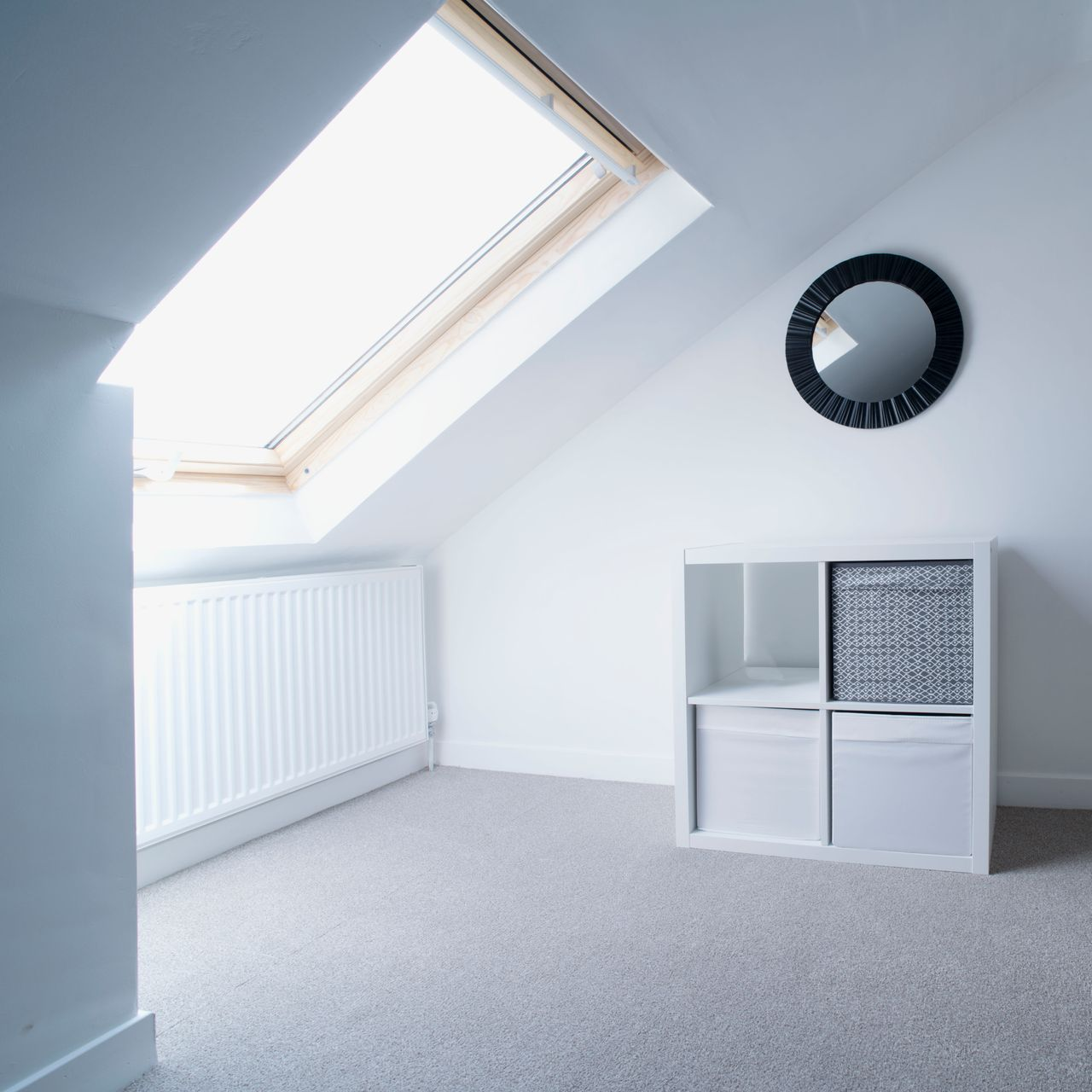 Chambre lumineuse avec plafond mansardé, puits de lumière, radiateur, étagère avec paniers et miroir rond.