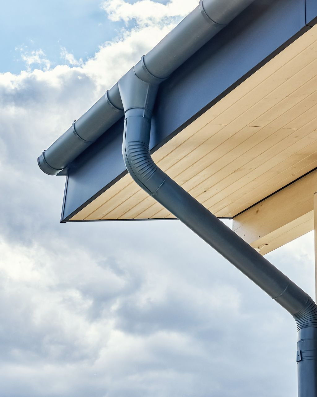 Gouttière grise sur une maison au toit bleu et aux avant-toits en bois. Ciel nuageux en arrière-plan.