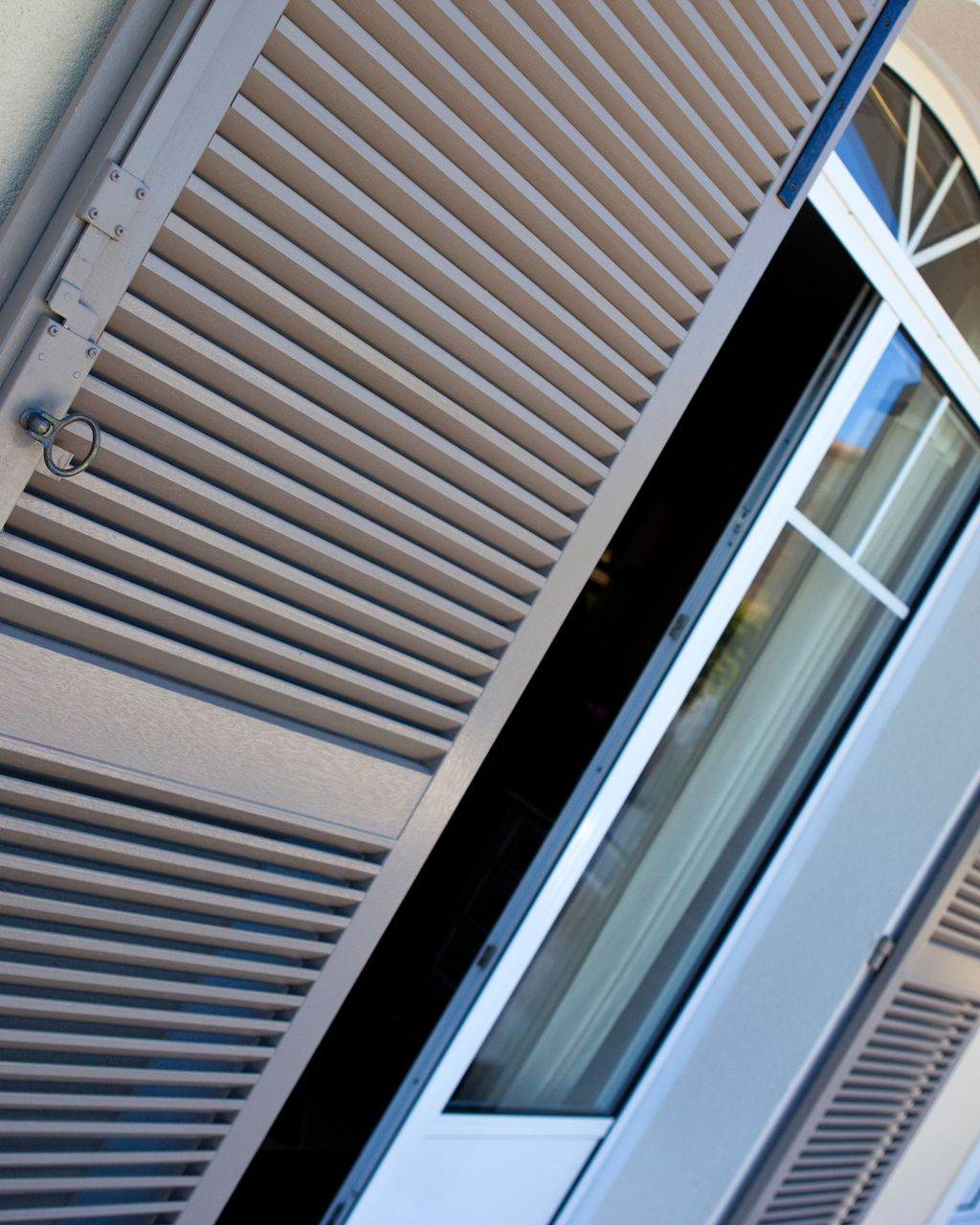 Fenêtre extérieure avec volets gris à persiennes partiellement ouverts. On aperçoit une fenêtre cintrée derrière.