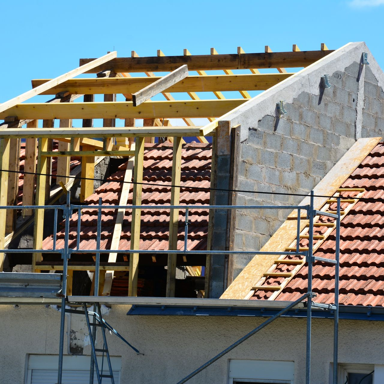 Toiture de maison en construction ; poutres en bois, blocs de béton, tuiles rouges, échafaudage.