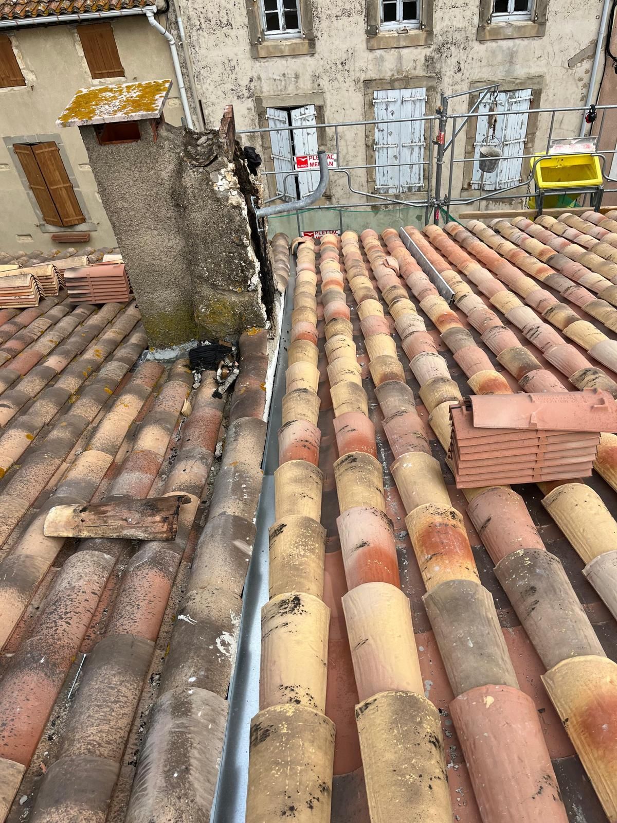 Toit en tuiles de terre cuite, cheminée et gouttière métallique. L'arrière-plan montre un bâtiment avec des fenêtres.