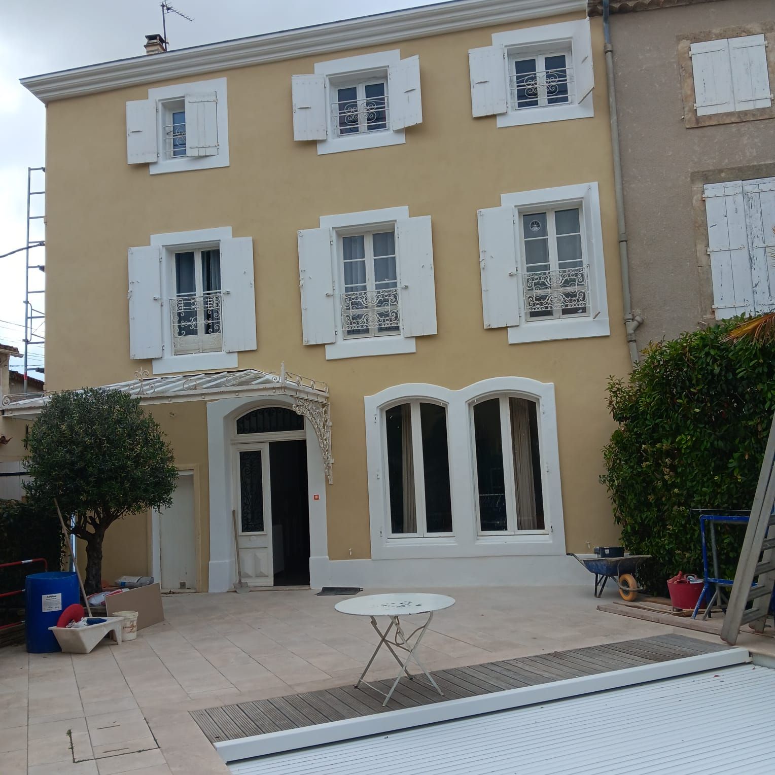 Maison jaune avec volets blancs et porte cintrée ; patio avant avec une petite table.