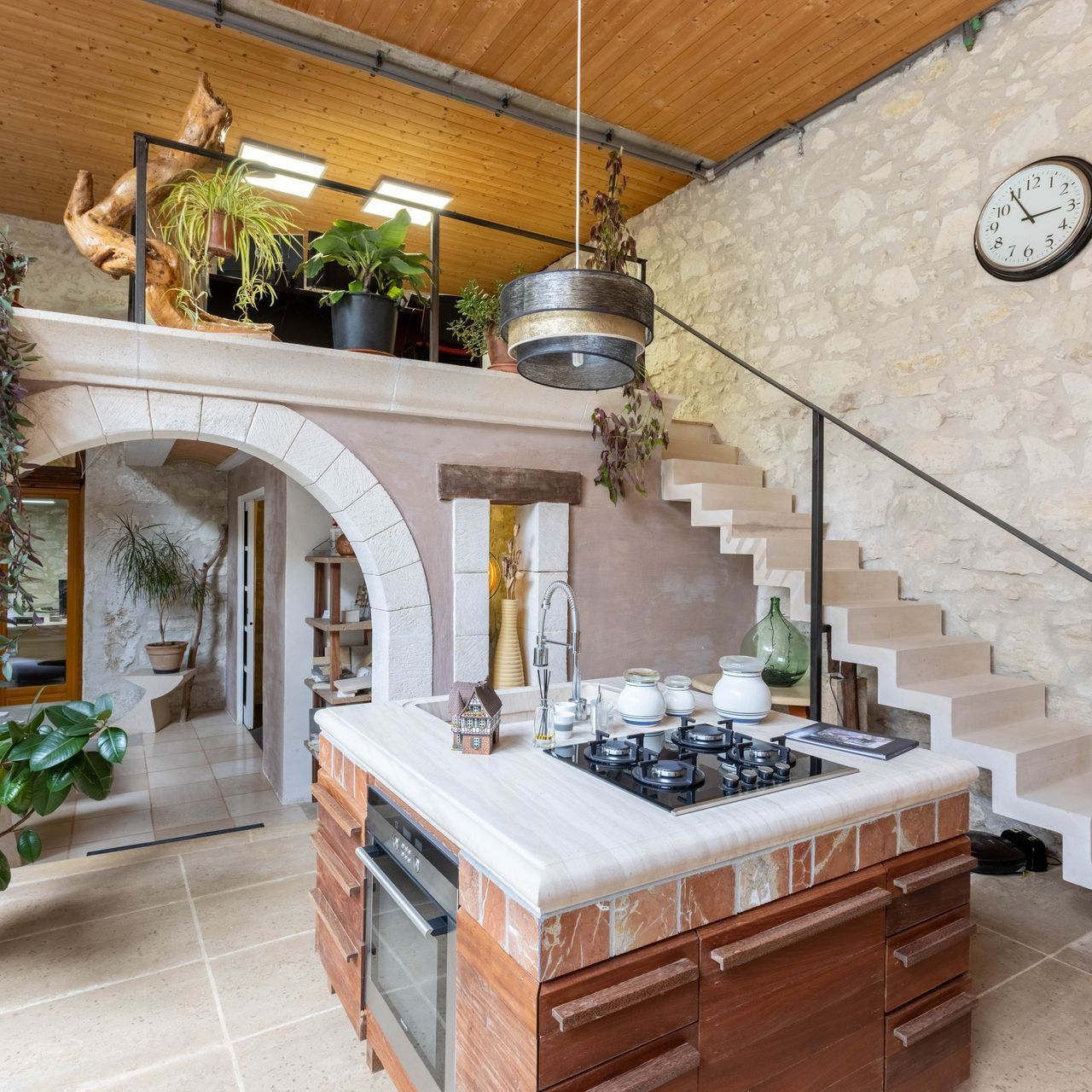 Cuisine avec mur en pierre, îlot central, mezzanine avec plantes et escalier en colimaçon.