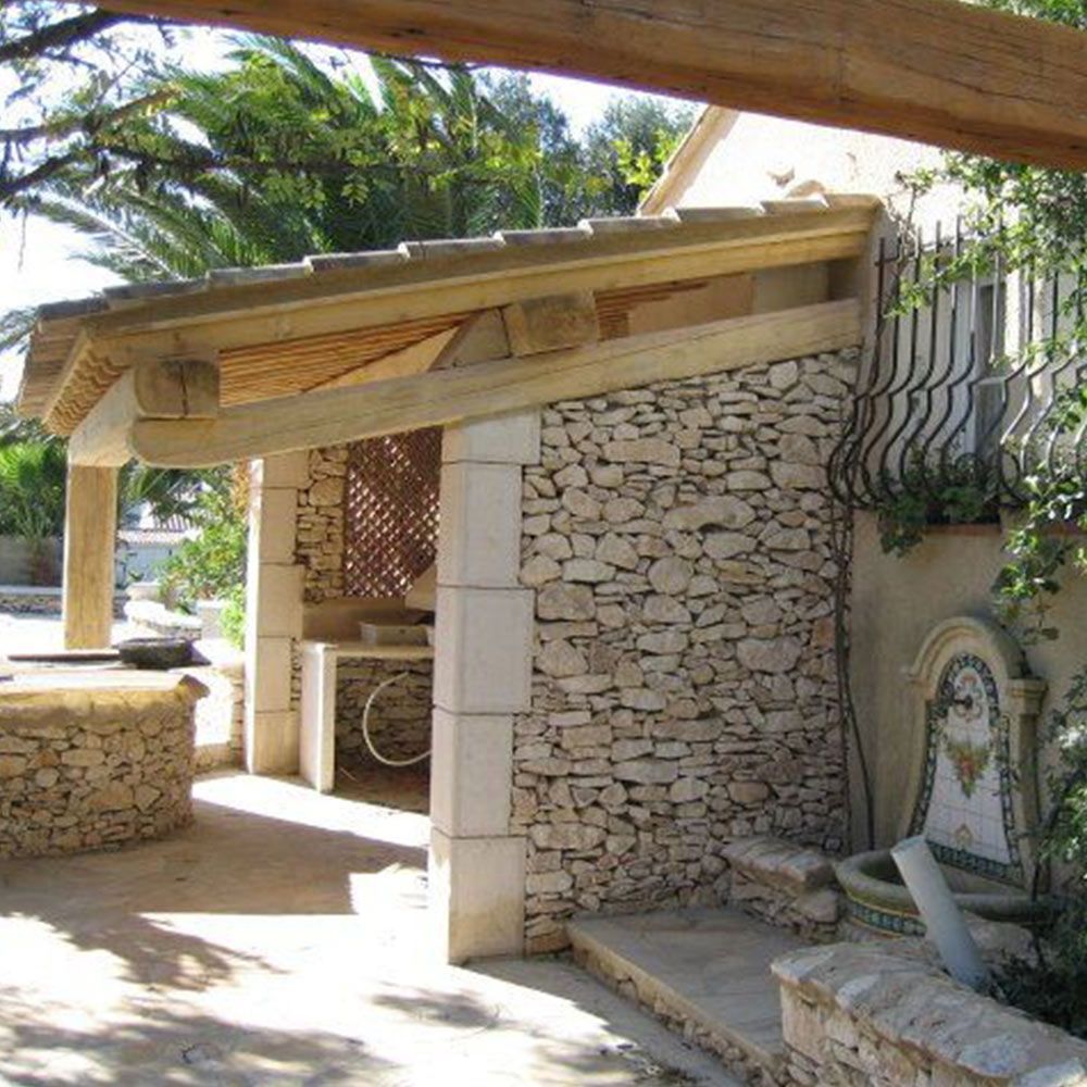 Cuisine extérieure avec murs en pierre, poutres en bois et toit en tuiles. Une fontaine décorative se trouve à droite.
