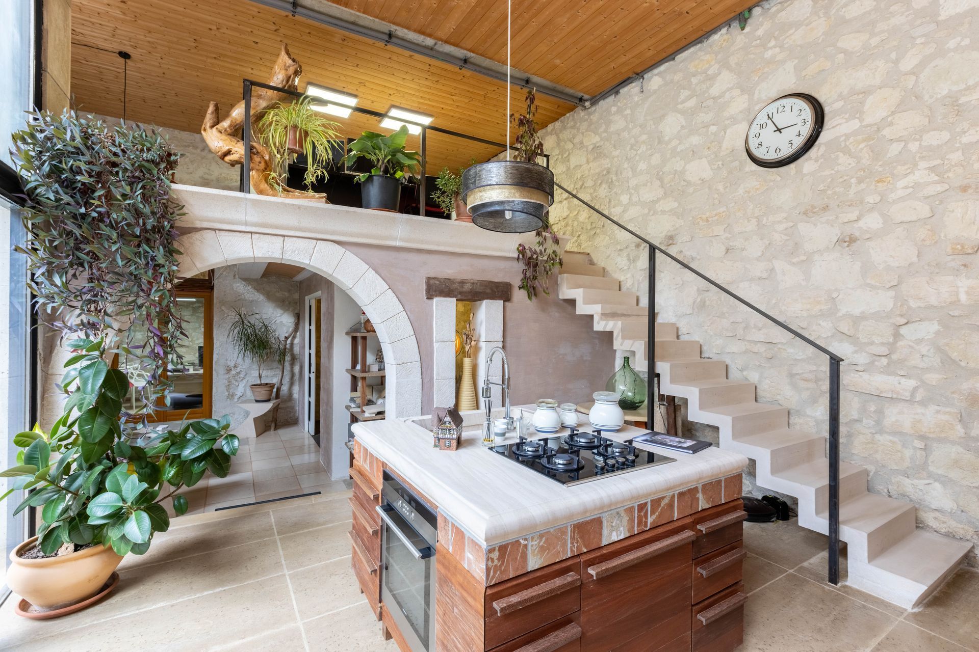 Cuisine avec mezzanine, murs en pierre et escalier. Elle dispose d'un îlot central avec cuisinière et plantes.
