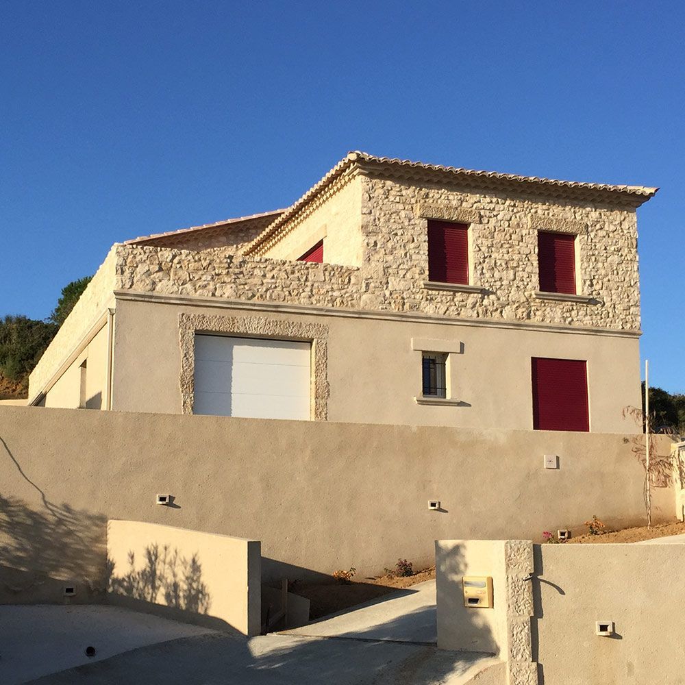 Maison en pierre et stuc aux volets rouges, posée à flanc de colline sous un ciel bleu.