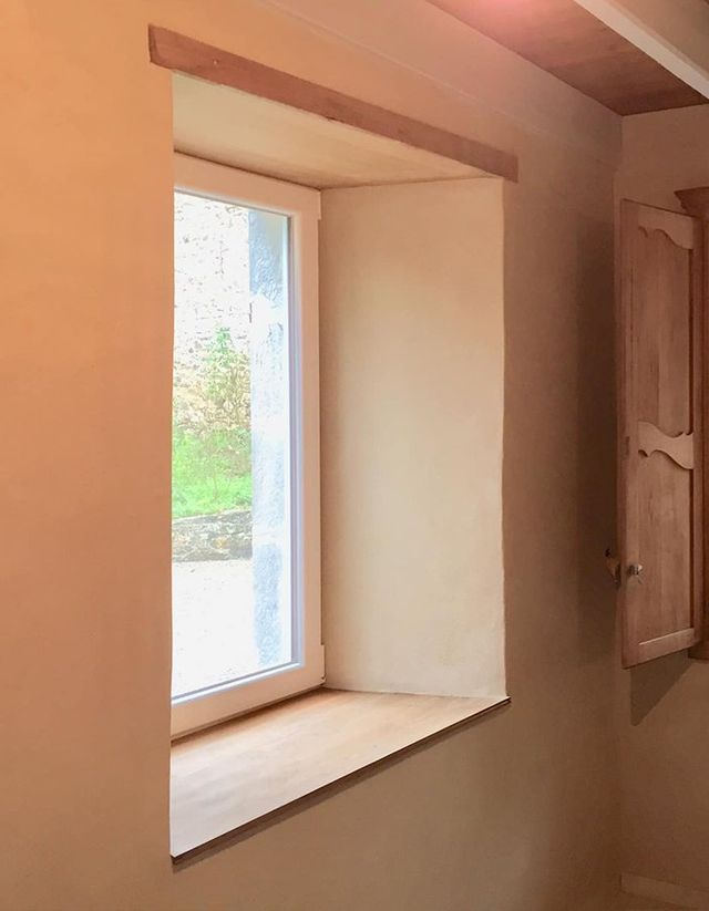 Fenêtre dans une pièce aux murs beiges avec boiseries et volets. Dehors, un jardin verdoyant.