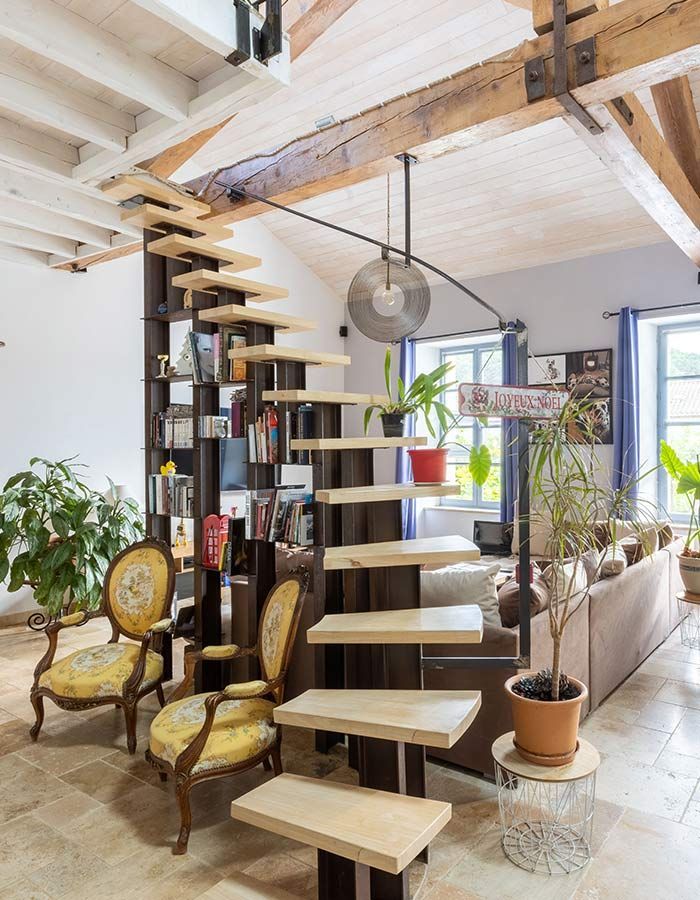 Escalier flottant en bois avec bibliothèque, chaises et plantes dans un salon lumineux et ouvert.