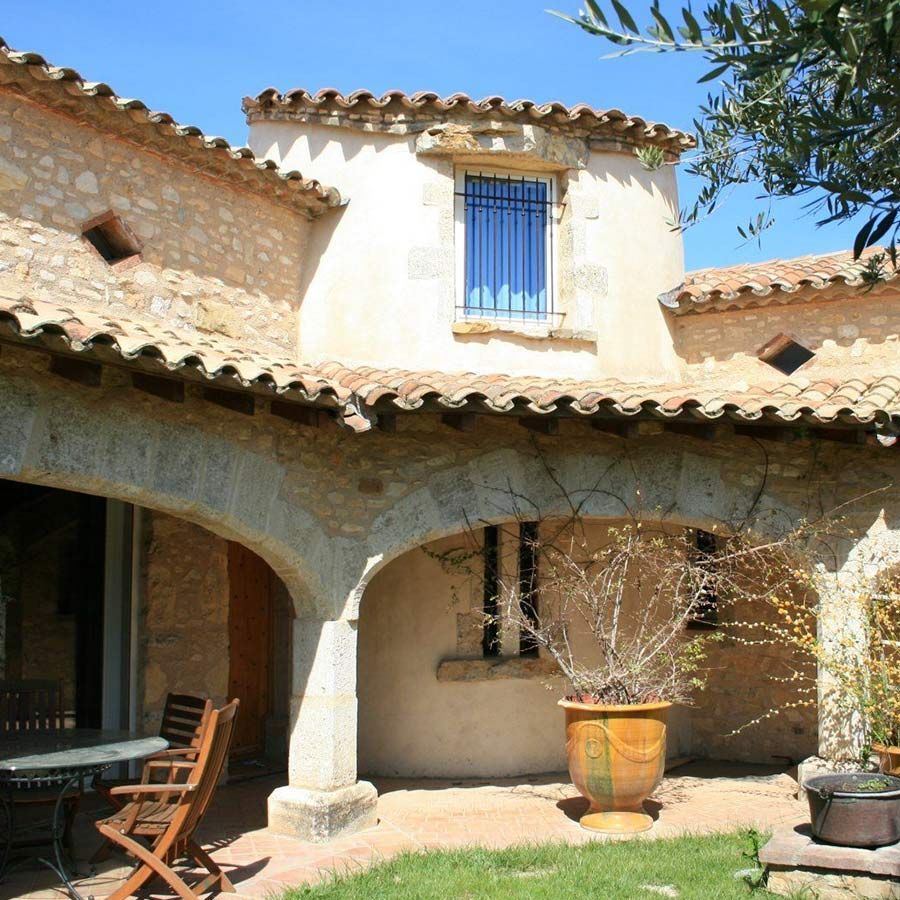 Bâtiment en pierre avec porche voûté, toit en terre cuite, fenêtre à volets bleus et plante en pot.
