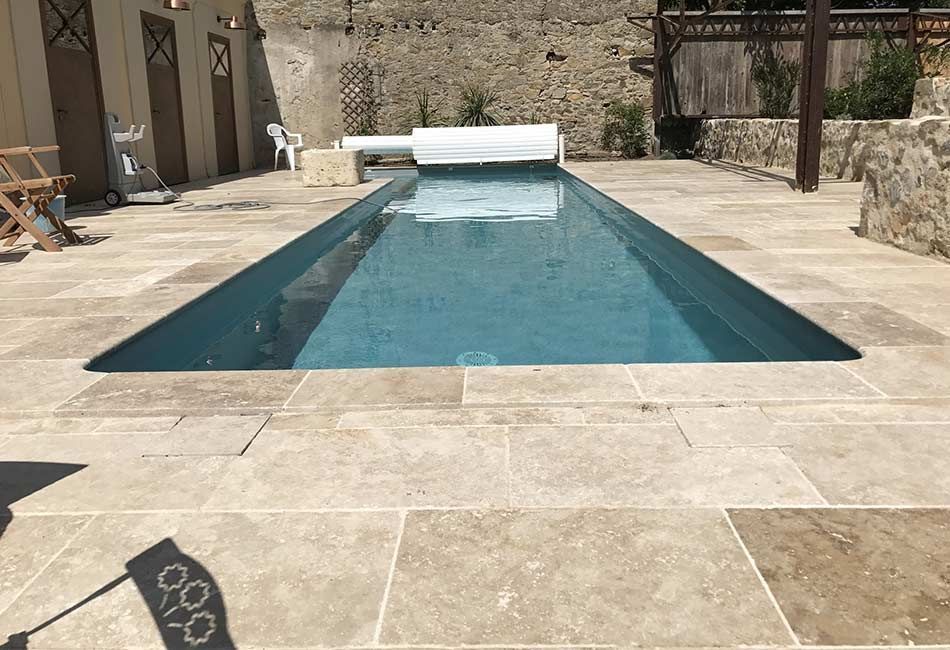 Piscine rectangulaire entourée d'un patio en pierre beige. Aménagement extérieur avec un mur de pierre en arrière-plan.