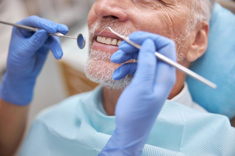 Ein Zahnarzt untersucht die Zähne eines älteren Patienten mit Mundspiegel und Sonde und trägt dabei blaue Handschuhe.