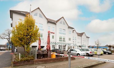 Weißes Hotelgebäude mit geparkten Autos, einem Baum und Sitzgelegenheiten im Freien unter einem bewölkten Himmel