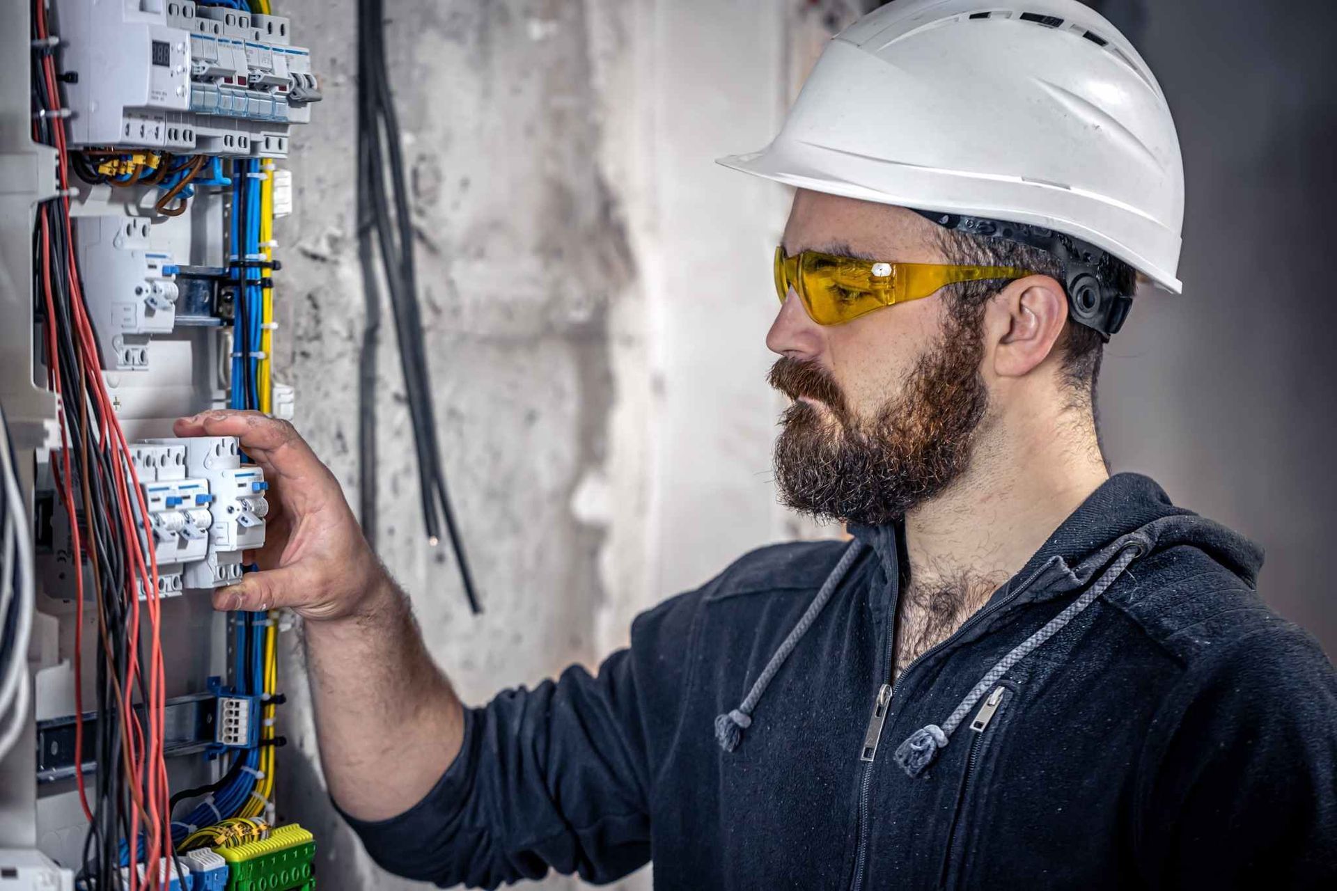 Electricista con casco y gafas de seguridad inspeccionando un panel eléctrico abierto en el interior de una vivienda.