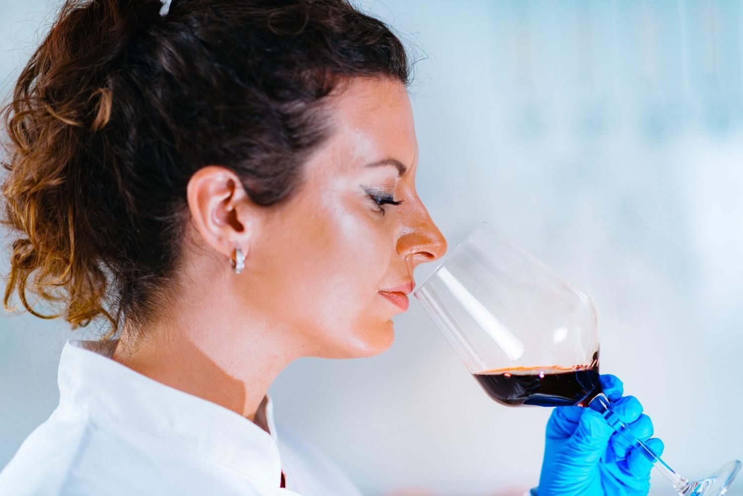 Una persona con bata blanca de laboratorio y guantes azules huele una copa de vino tinto en un laboratorio.