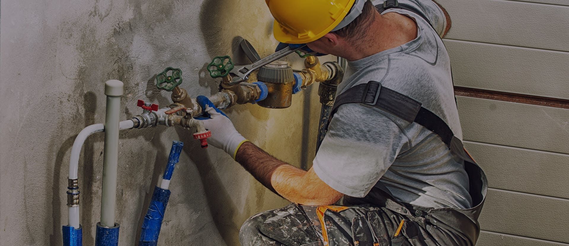 Un ouvrier portant un casque de chantier jaune resserre des tuyaux à l'aide d'outils sur un système de plomberie mural.