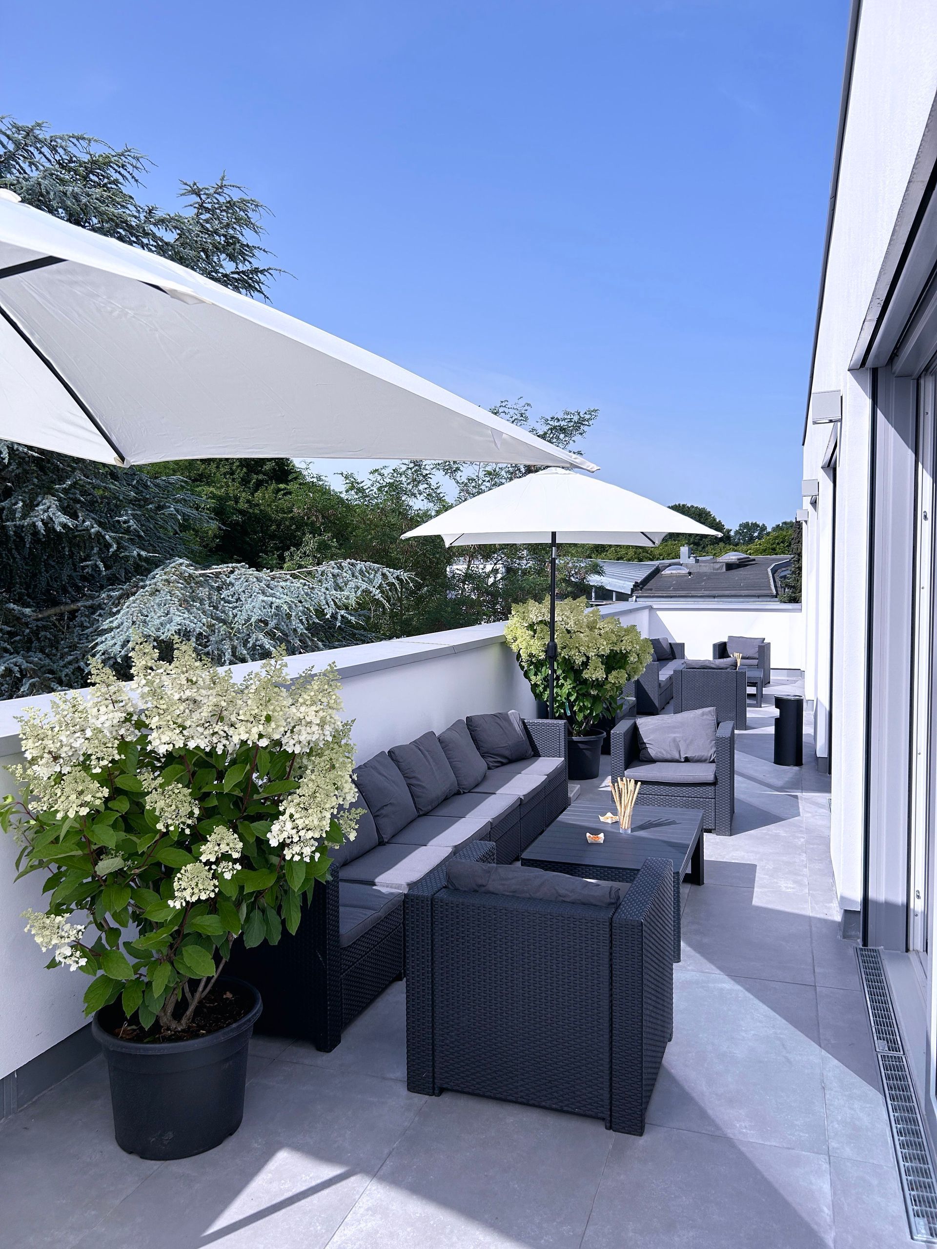 Terrasse mit dunkelgrauen Möbeln, weißen Sonnenschirmen und Topfblumen an einem sonnigen Tag.