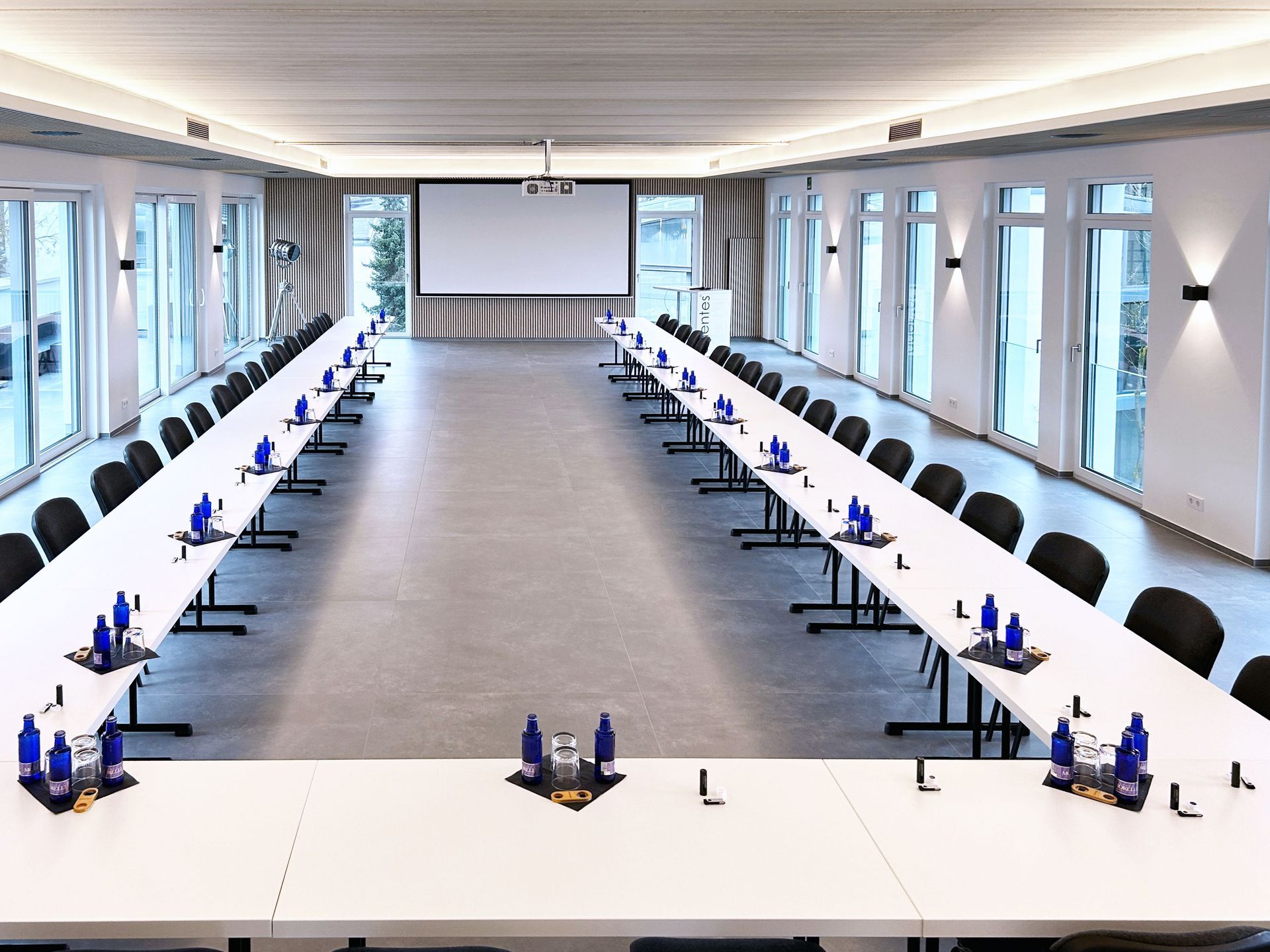 Konferenzraum mit langen, weißen Tischen in U-Form, mit Wasserflaschen gedeckt, Fenster an den Wänden.