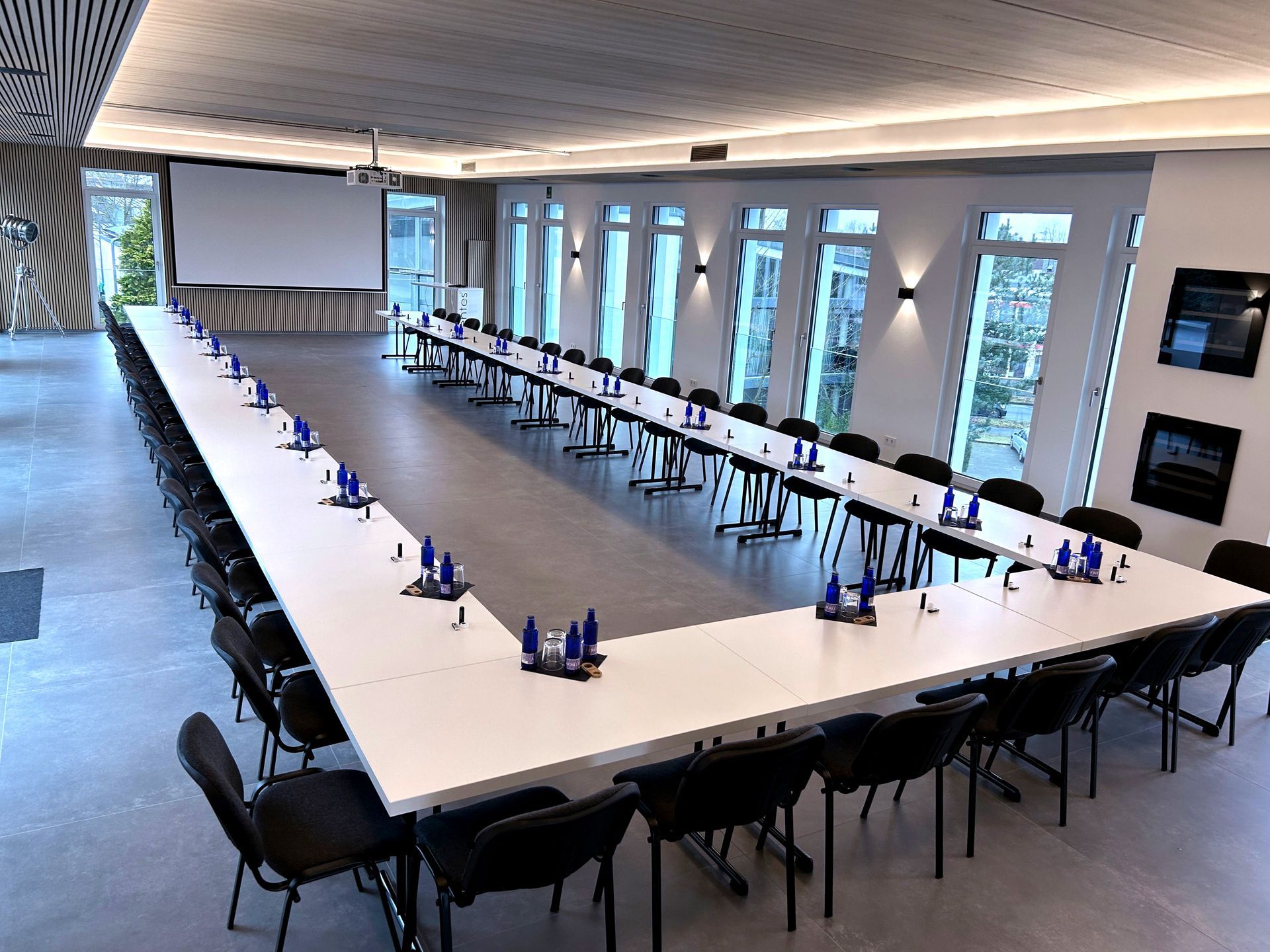 Konferenzraum mit U-förmigem Tisch, schwarzen Stühlen, Projektionsleinwand und Fenstern.