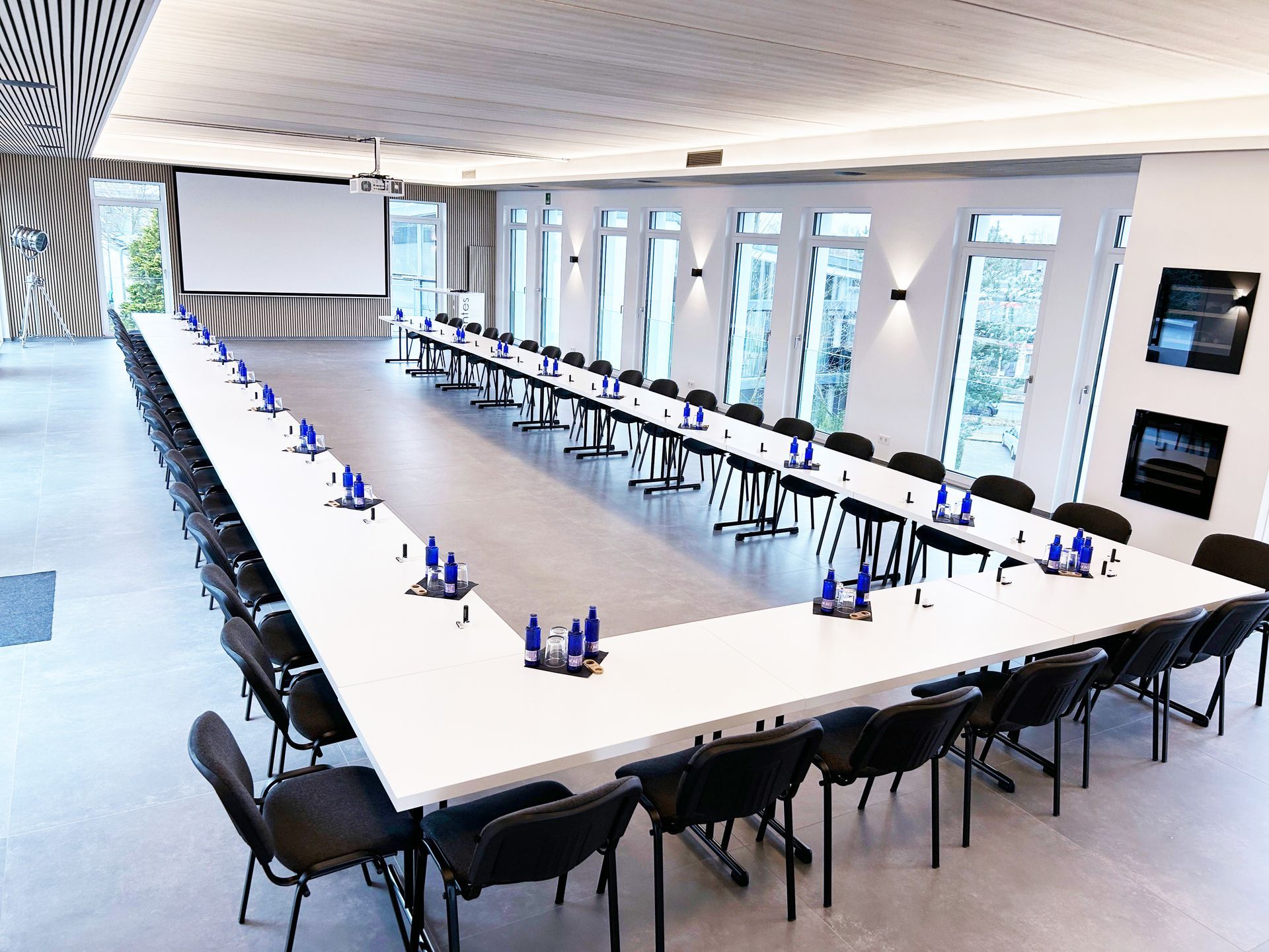 Ein U-förmiger Konferenztisch mit Stühlen in einem hellen, modernen Raum mit großen Fenstern und einer Projektionsfläche.