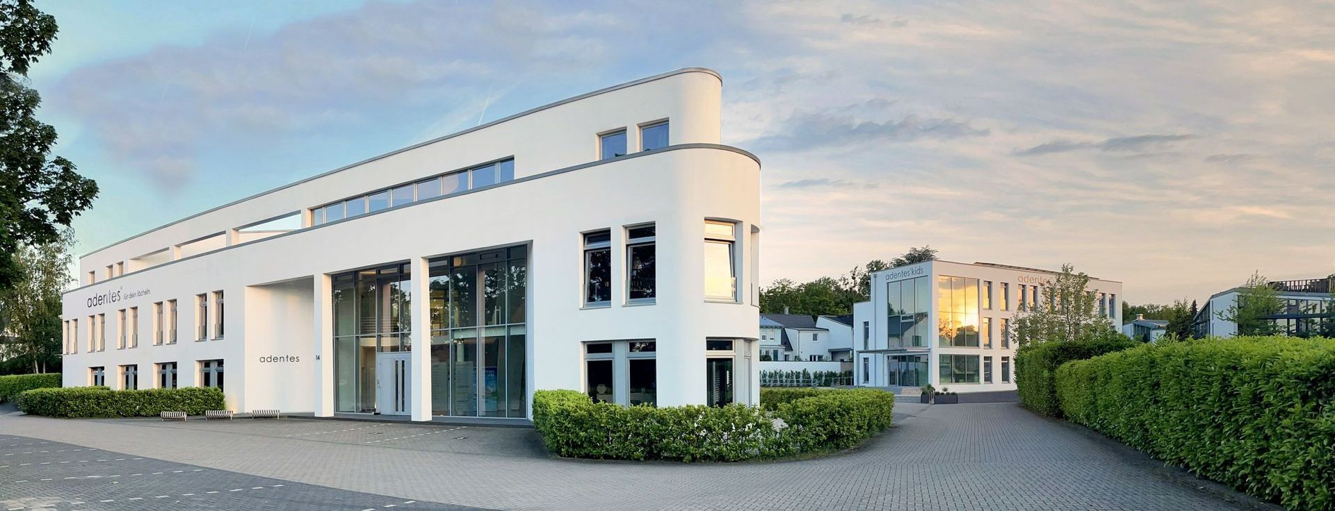 Modernes weißes Gebäude mit abgerundeter Ecke und Glaseingang, umgeben von grünen Büschen und einer gepflasterten Auffahrt.