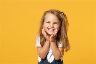Kinderzahnheilkunde Ein kleines Mädchen lächelt und schlägt die Hände vors Gesicht.