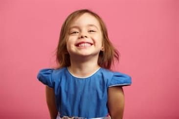Kinderzahnheilkunde Ein kleines Mädchen in einem blauen Kleid lächelt vor einem rosa Hintergrund.