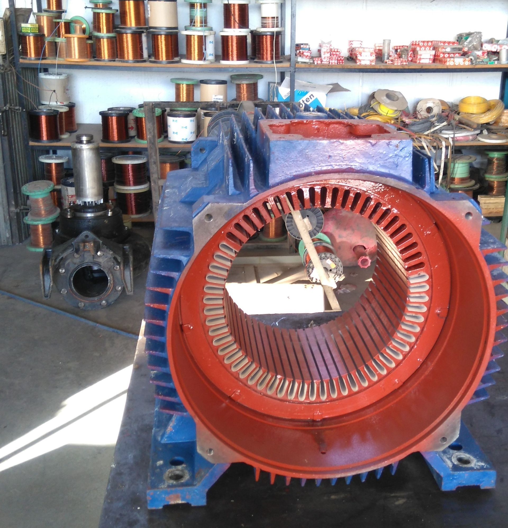 Estátor de motor eléctrico grande, rojo y azul, con carretes de alambre de cobre al fondo. Ambiente de taller.