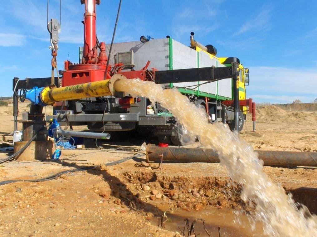 Operación de perforación de pozo; agua marrón brota a borbotones de una tubería amarilla hacia una zanja; camión grande con grúa en un entorno desértico.