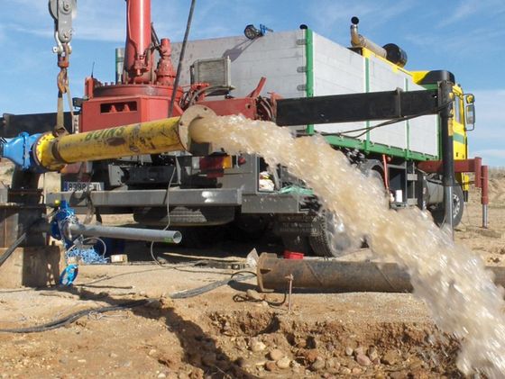 Agua brotando a borbotones de una tubería conectada a una plataforma de perforación; paisaje desértico de color beige.