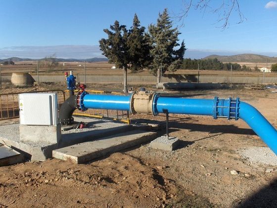 Tuberías de agua azules con válvulas y caja de control, en un entorno exterior con campos y colinas.