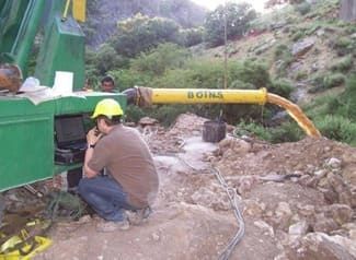 Hombre con casco trabajando cerca de una plataforma de perforación mientras un líquido marrón fluye de una tubería.