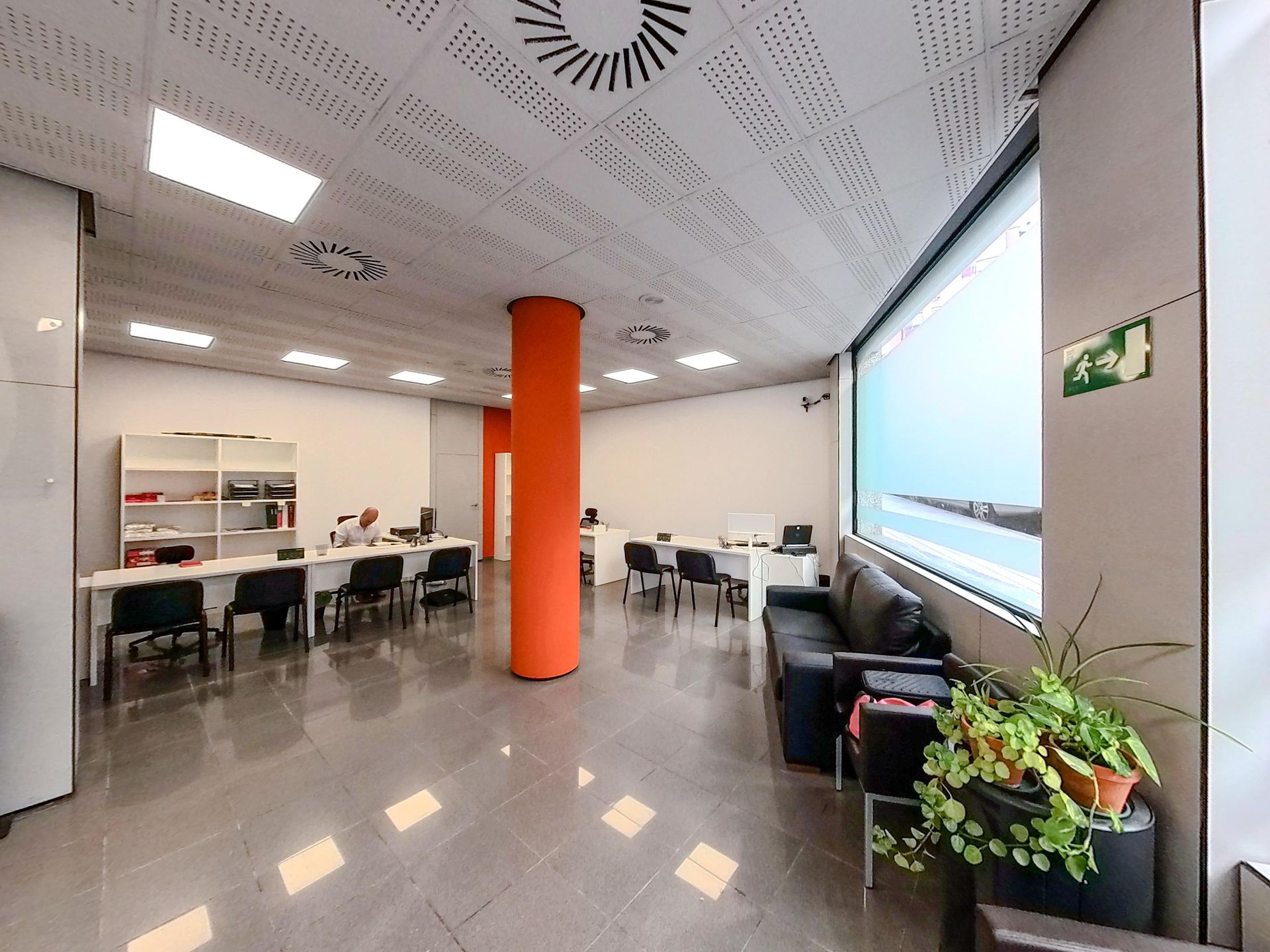 Moderno salón de oficina con escritorios, sillas, una columna naranja y una amplia zona de asientos junto a la ventana