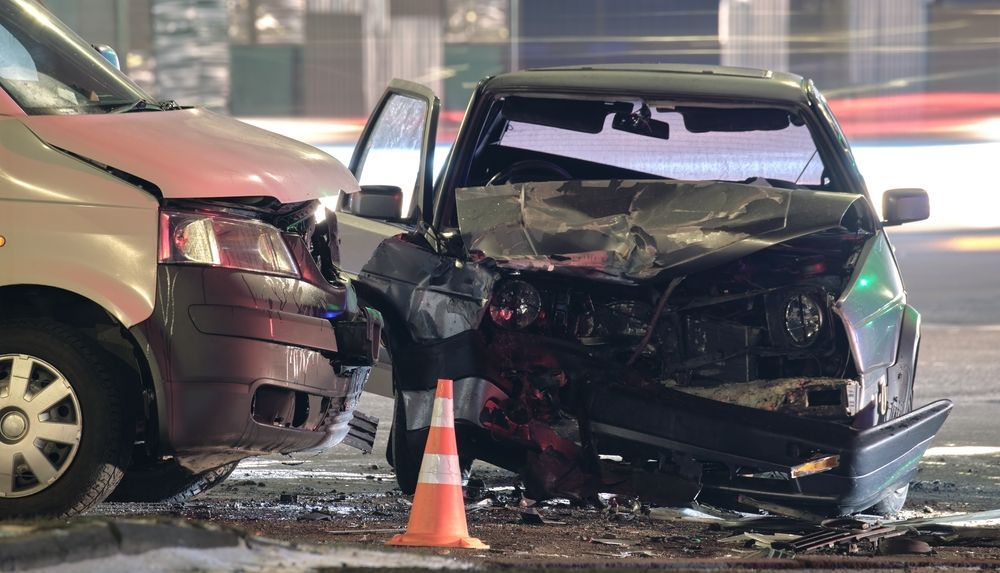 Grave choque entre dos coches por la noche, con los frontales destrozados alrededor de un cono naranja en una calle mojada.