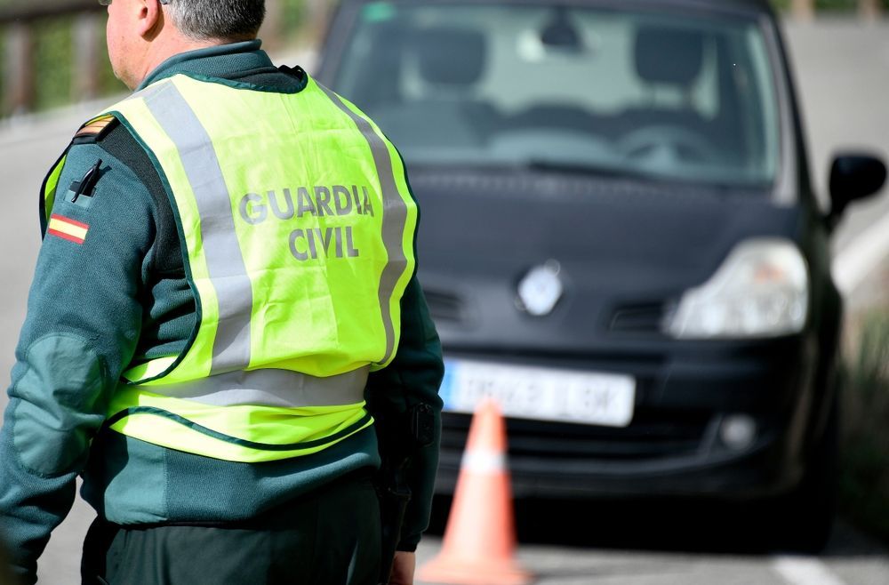 Un agente de la Guardia Civil con un chaleco reflectante dirige el tráfico junto a un cono naranja, con un coche negro detrás