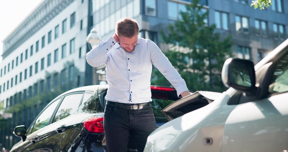 Hombre de pie junto a un coche blanco dañado en una calle de la ciudad, sujetándose la cabeza con frustración