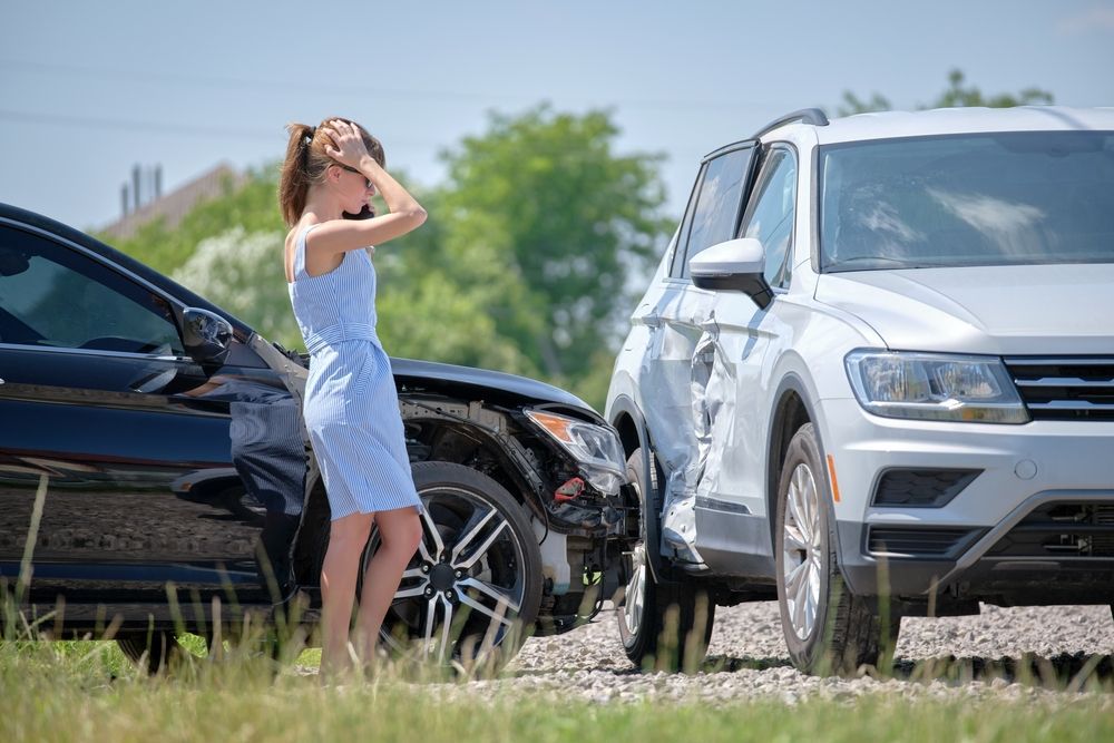 Una mujer de pie junto a dos coches dañados al borde de la carretera, protegiéndose los ojos del sol intenso.