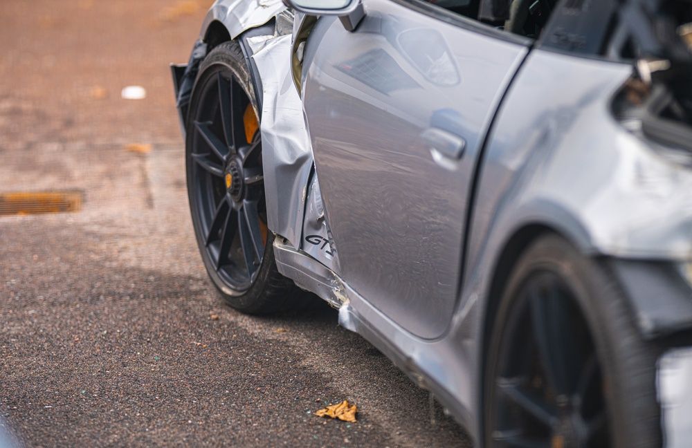 Coche deportivo plateado dañado, con un lateral rozado y la rueda trasera doblada, sobre el pavimento