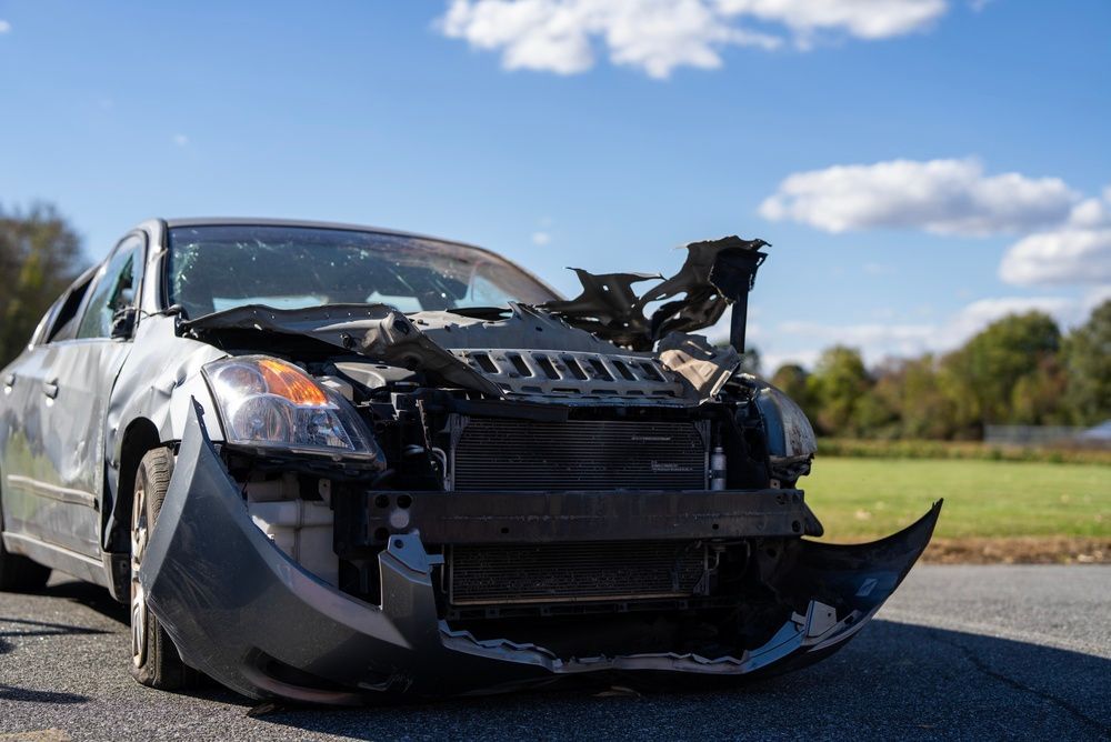 Un coche negro gravemente dañado a un lado de la carretera, con la parte delantera destrozada y el parachoques colgando bajo un cielo azul