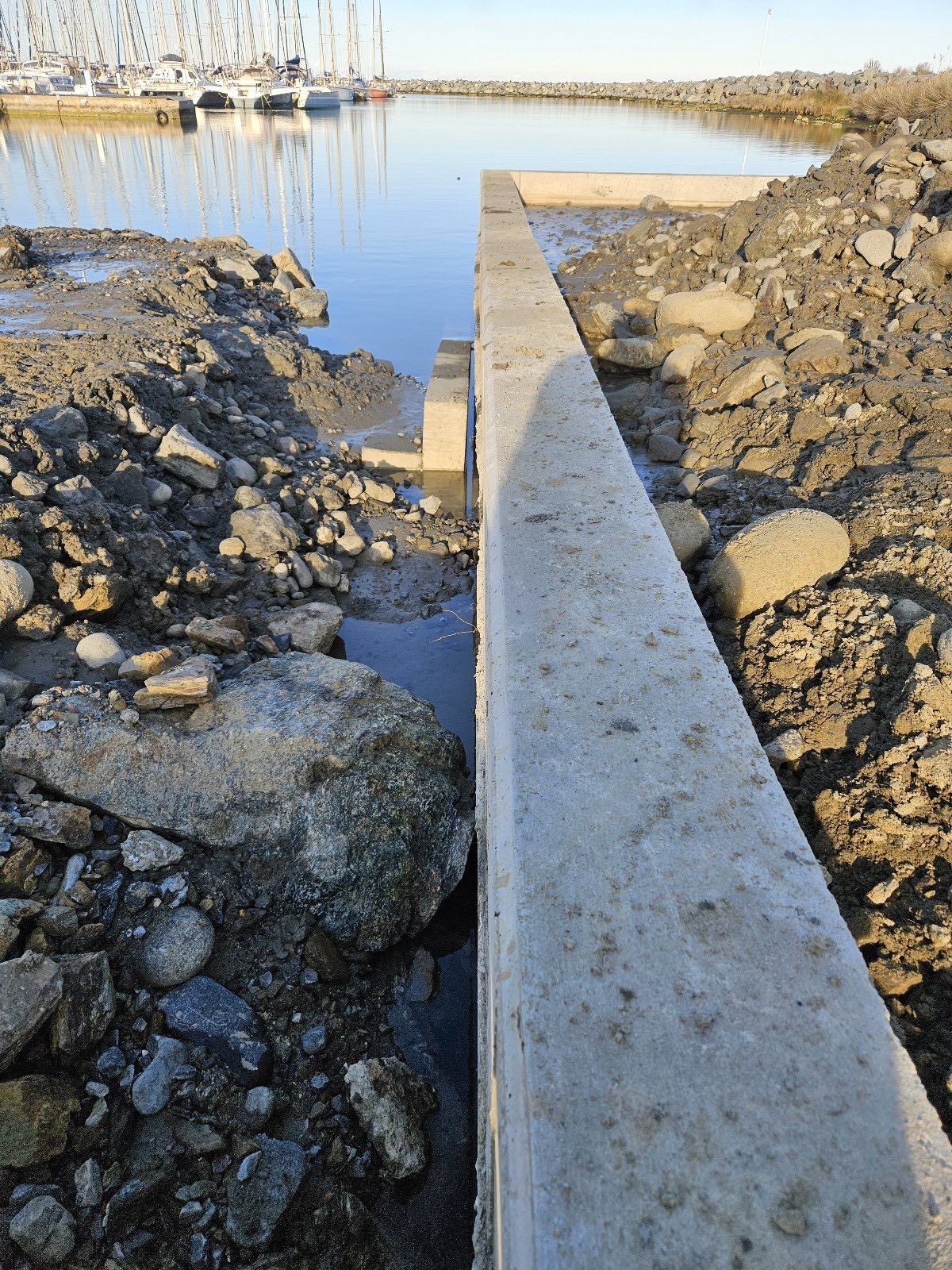 Barrière en béton séparant la boue et l'eau ; bateaux dans le port de plaisance en arrière-plan