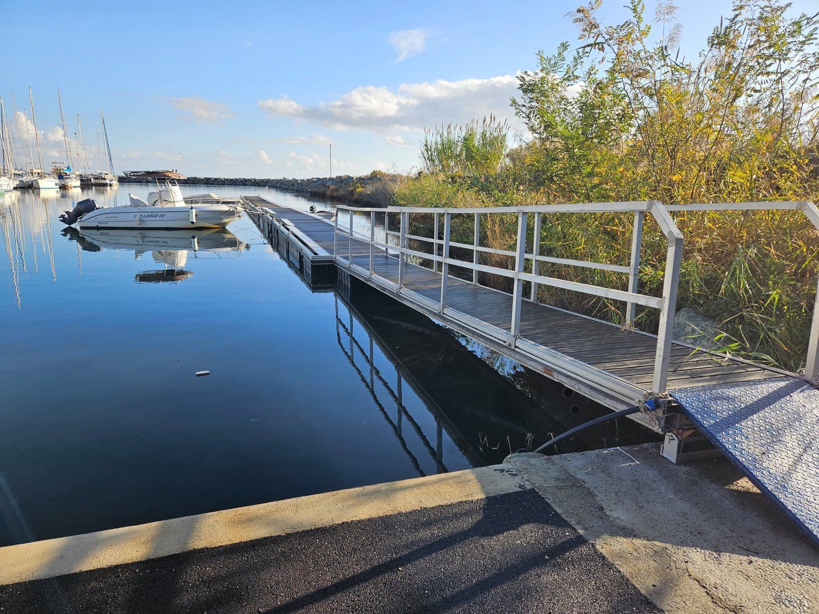 Une rampe métallique mène à un quai qui s'avance dans un plan d'eau où se trouvent des bateaux