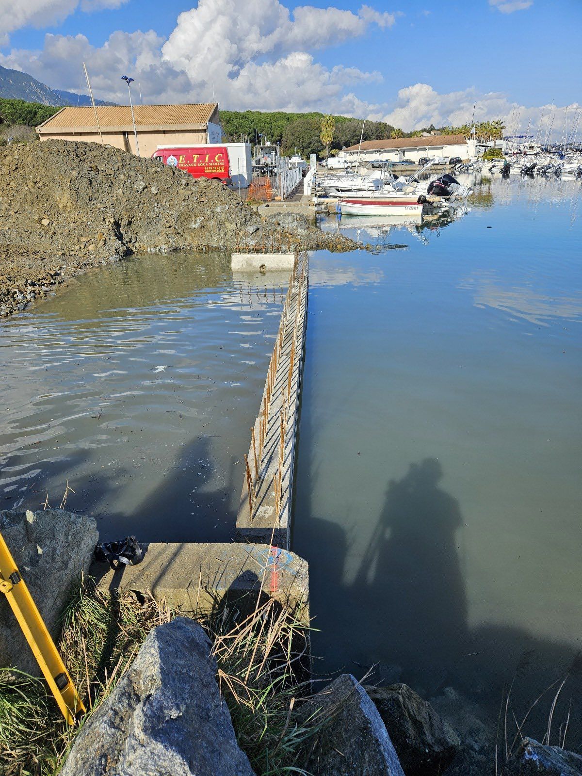 Travaux en bord de mer ; une barrière en béton s’avance dans l’eau près de bateaux amarrés et de bâtiments