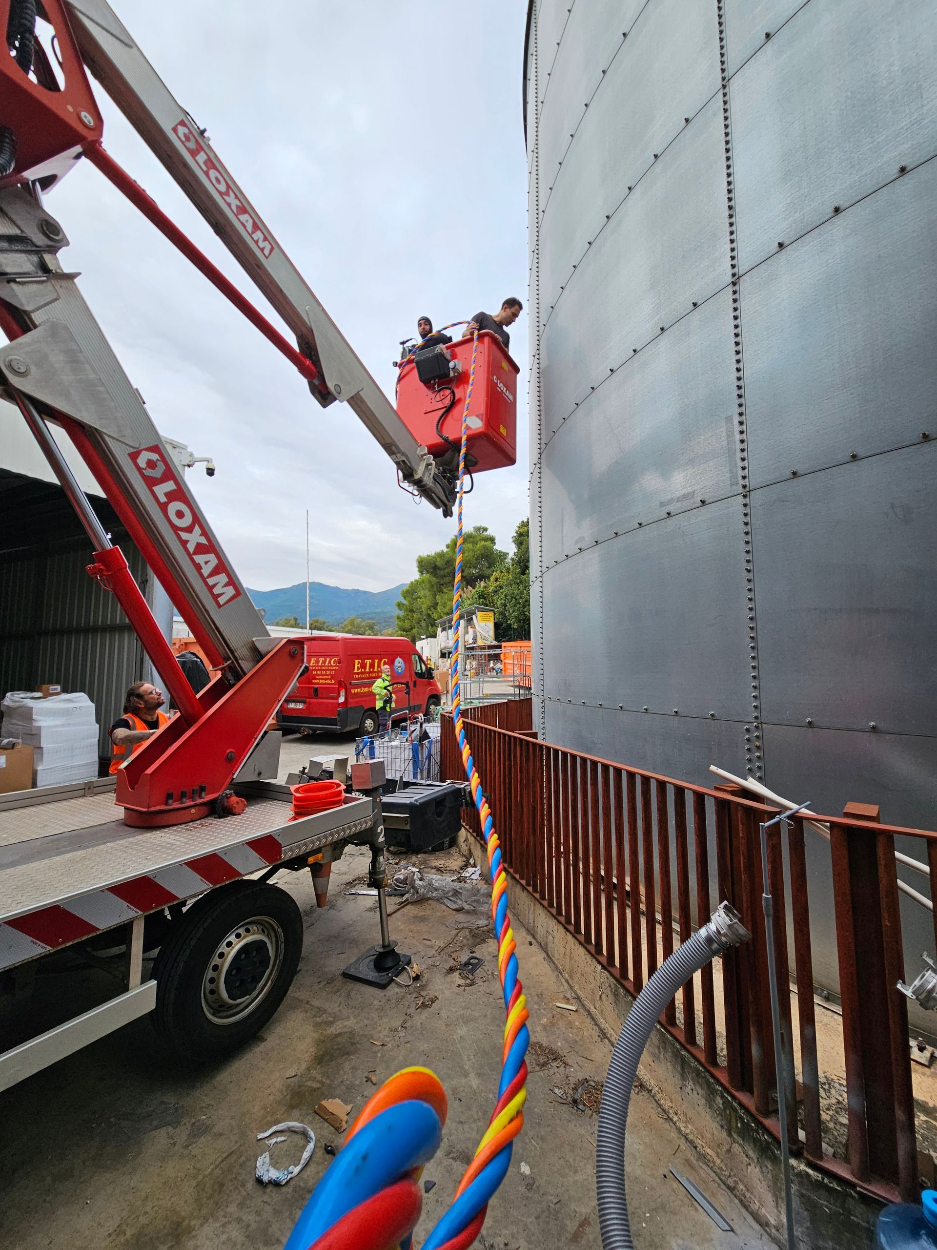 Des ouvriers dans une nacelle élévatrice rouge près d'un grand réservoir métallique