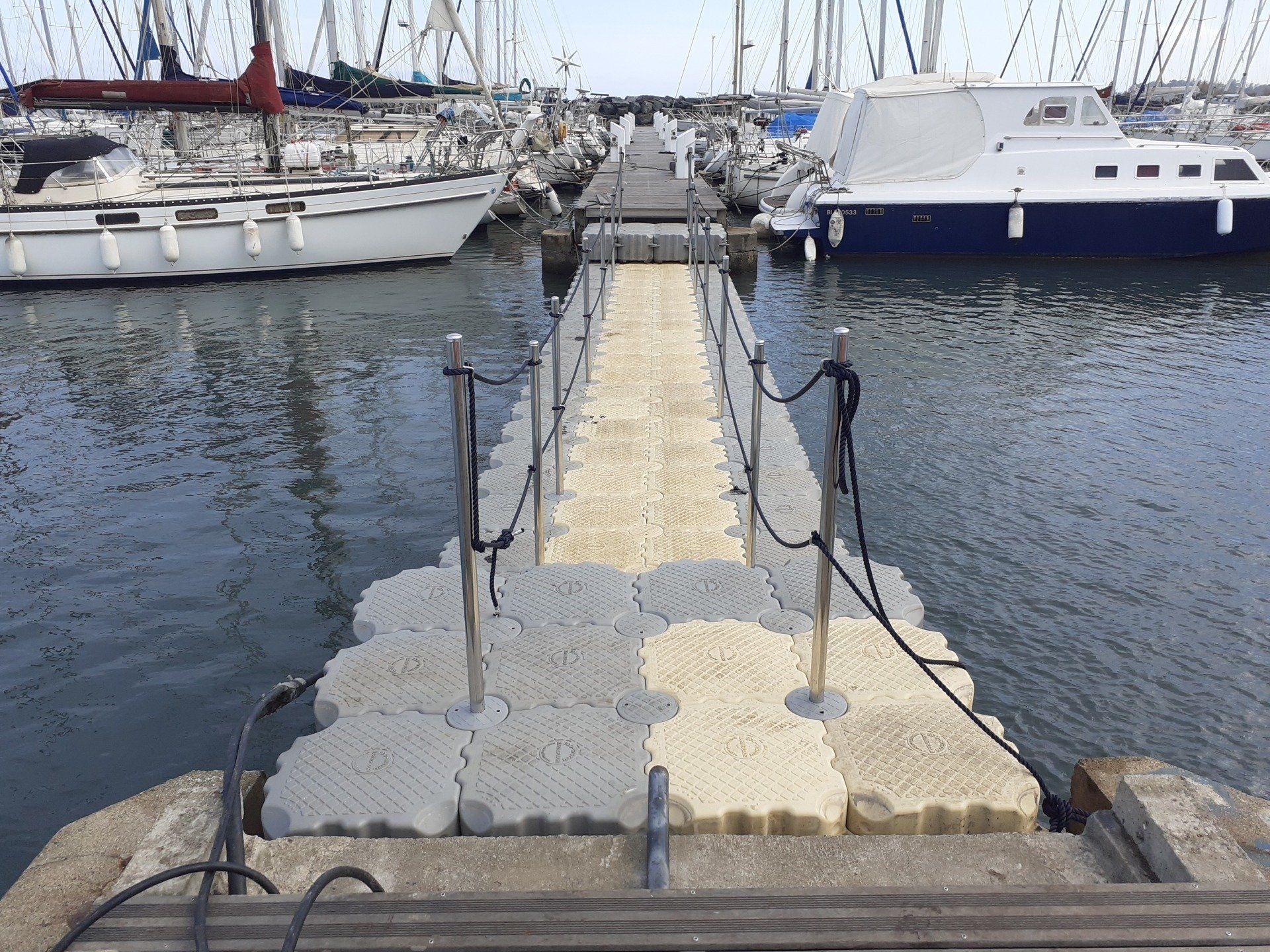 Quai flottant d'une marina, menant à des voiliers amarrés. Eaux grises, journée ensoleillée