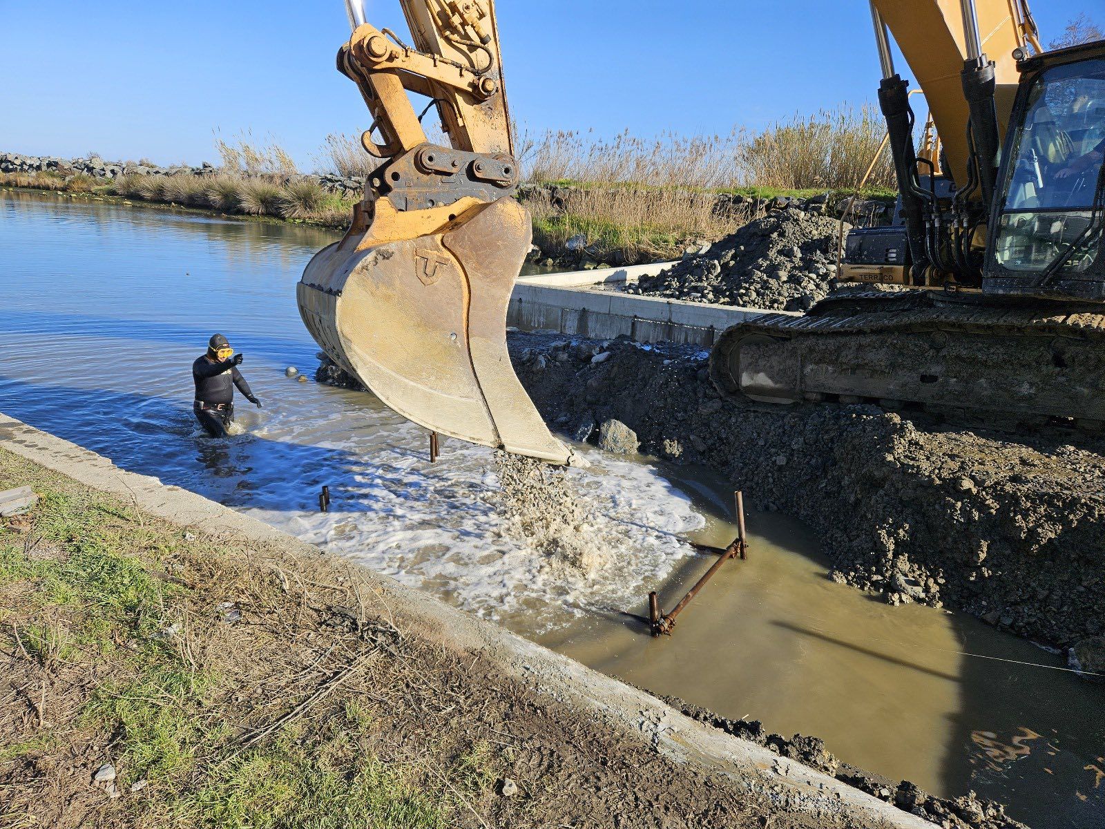 Une excavatrice déverse des matériaux dans un canal, tandis qu'une personne en cuissardes se tient dans l'eau