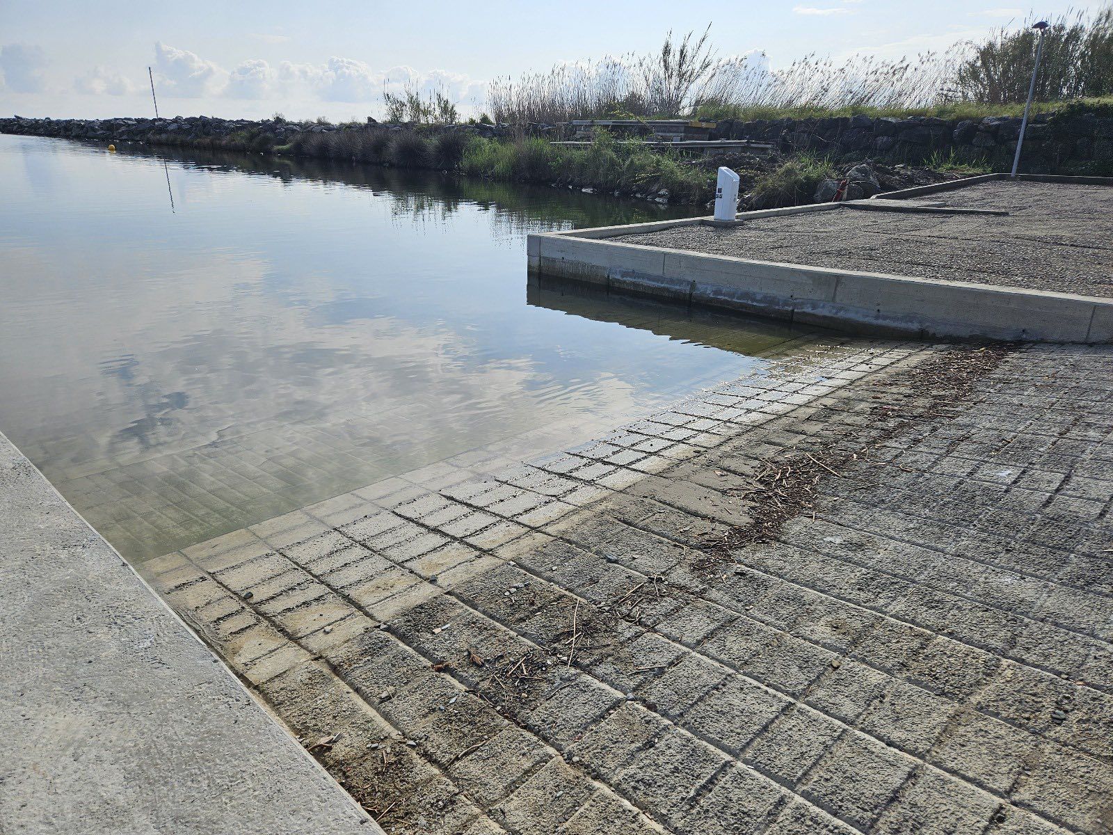 Cale de mise à l'eau en béton donnant sur des eaux calmes et claires, avec des roseaux et un talus de pierre
